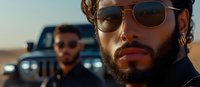 Two men wearing sunglasses stand near vehicles in a desert landscape. The foreground features a bearded male with reddish-brown hair and facial hair, appearing to be of mixed ethnicity, wearing dark sunglasses and casual attire. A second male is visible in the background near a vehicle. The scene suggests an outdoor adventure or road trip setting.