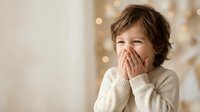 Young child with curly hair covering mouth with hands in cozy cream sweater against warm bokeh lights background for holiday family moments.