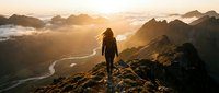 Female hiker silhouette standing on mountain peak at golden sunrise overlooking dramatic valley landscape with winding river and misty peaks below.