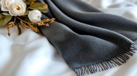 Luxurious gray cashmere scarf with fringe detail on white silk background, decorated with cream roses and golden ornamental elements. Elegant fashion composition.