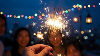 Hands holding lit sparkler at night with blurred bokeh lights and people in background, suitable for celebration, party, and festive event marketing materials.