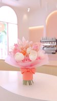 Delicate pink and white floral bouquet wrapped in soft pink paper with bow accent, featuring roses and tulips on light counter in pastel cafe interior setting.