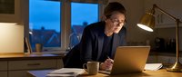 Mature Caucasian businesswoman working late at home office, focused on laptop screen by warm desk lamp at night.