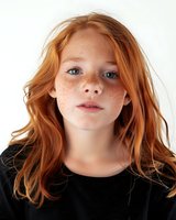 Young redhead girl with natural freckles and bright blue eyes wearing black top against white background, looking directly at camera with gentle expression.