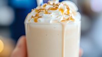 Creamy caramel latte topped with whipped cream and drizzled with golden caramel sauce, served in clear glass against blurred lights background.