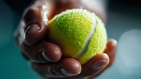 Hand holding bright yellow tennis ball with water droplets against dark background, dramatic lighting creates atmospheric sports composition.