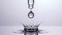 Water drop falling creating splash crown with ripples on clean surface against soft purple background for wellness and purity concepts.