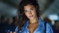 Young woman with wavy dark hair wearing blue striped shirt and gold necklace in soft focus indoor setting with dramatic lighting.