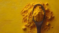 Ground turmeric powder scattered on yellow background with wooden spoon, overhead view showing vibrant golden spice texture and natural cooking ingredient.