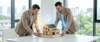 Two middle-aged Caucasian male professionals in business casual attire examining a wooden architectural model on a white table in a modern high-rise office with city skyline views, representing real estate development and collaboration.