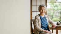 Senior woman with gray hair and East Asian ethnicity, wearing casual cardigan and blue top, sitting by window in bright home interior with natural light and potted plant.
