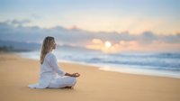 Young blonde woman in white clothing practicing meditation yoga on sandy beach at sunrise with peaceful ocean waves and golden sky creating serene wellness atmosphere.