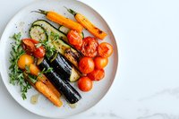 Grilled seasonal vegetables on white plate with fresh herbs and olive oil dressing, featuring colorful carrots, zucchini, eggplant and cherry tomatoes on marble surface.
