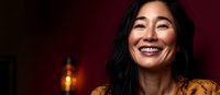 Cheerful Asian woman with genuine smile wearing yellow floral dress against dark red background with warm ambient lighting, expressing joy and confidence.