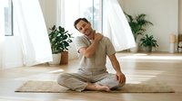 Young Caucasian man sitting cross-legged on yoga mat, stretching shoulder, smiling in bright minimalist studio with plants.
