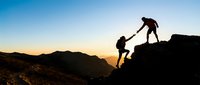 Two hikers helping each other climb rocky mountain terrain during golden hour sunset with dramatic sky and mountain silhouettes in background.