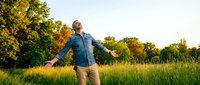 Young man with arms outstretched enjoying freedom in sunny meadow with golden hour lighting and lush green trees for wellness and lifestyle concepts.