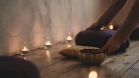 Relaxing spa meditation scene with singing bowl, candles and crossed legs on wooden floor creating peaceful wellness atmosphere for mindfulness practice.
