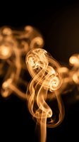 Golden smoke swirls curl elegantly against a dark black background, creating abstract flowing shapes with warm amber tones and soft bokeh effect.