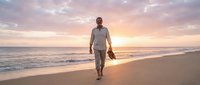 Middle-aged man with dark skin tone wearing light linen shirt and pants walking barefoot on sandy beach at sunset, carrying shoes, ocean waves in background, golden hour lighting.