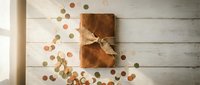 Rustic gift box wrapped in brown paper with twine bow surrounded by scattered golden confetti on white wooden background for celebrations.