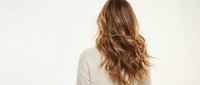 Young woman with long wavy brown hair from behind wearing cream sweater against neutral background for hair care and beauty concepts.