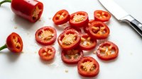 Red bell pepper sliced into rings with seeds visible, arranged on white surface with whole pepper and knife for cooking preparation.