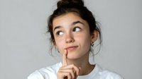 Young woman with messy bun hairstyle making thoughtful expression with finger on lips against light background, looking up with playful contemplative gaze.