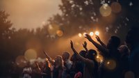 Crowd of people raising hands at outdoor music festival during golden hour sunset with warm bokeh lights and silhouetted trees creating atmospheric concert scene.