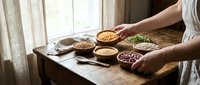 Hands holding bowls of assorted legumes, grains and seeds on wooden table by window for cooking and nutrition preparation.
