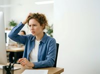 Caucasian woman with curly reddish-brown hair, wearing blue shirt over white top, sitting at desk in modern office, hand raised to head in thoughtful or stressed pose, working environment with plants and natural light.