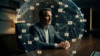 Middle-aged man with dark hair and facial hair, appearing to be of South Asian or Mediterranean ethnicity, wearing a dark blazer and light shirt, seated at a desk with hands clasped, surrounded by glowing email icons and a digital globe network visualization representing global communication and connectivity.