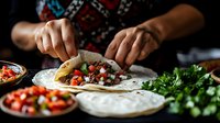 Young adult male of Middle Eastern or Mediterranean ethnicity preparing traditional tacos with fresh vegetables and herbs, hands actively assembling food on white tortillas with colorful ingredients visible in bowls nearby.