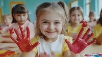 Happy blonde preschool girl showing red painted hands in art class with diverse children creating colorful artwork in bright educational setting.