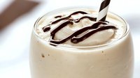 Creamy vanilla milkshake topped with rich chocolate syrup drizzle and decorative spoon in glass, perfect for dessert menus and beverage advertising.