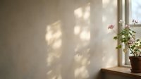 Sunlight streaming through window creates geometric shadows on beige wall beside potted pink flowers on wooden sill, ideal for interior design and home decor projects.