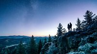 Silhouette of two people standing on rocky mountain peak at twilight with starry sky, forested valley below, and distant mountains, capturing adventure and exploration at dusk.