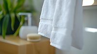 Clean white cotton towel hanging in bright bathroom with natural light, green plant and wooden accessories creating spa-like atmosphere for wellness content.