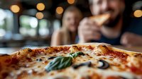 Close up of fresh baked Neapolitan pizza with melted mozzarella, olives and basil leaves on wooden table, with blurred couple dining in restaurant background.