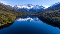 Aerial view of pristine mountain lake reflecting snow-capped peaks and surrounding wilderness, calm blue water surface mirroring majestic alpine landscape.