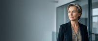 Middle-aged Caucasian woman in professional business attire, wearing dark blazer and gray blouse, standing in modern office environment with glass partitions and natural lighting, suitable for corporate, leadership, and business concepts.