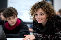 Caucasian female teacher with curly brown hair smiling while helping student with laptop in classroom setting. Professional education environment shows positive learning interaction.