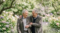 Two senior Caucasian men walking together in blooming spring garden, enjoying friendship and conversation among flowering trees and plants.