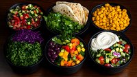Fresh Mediterranean food bowls with colorful vegetables, chickpeas, flatbread, and tzatziki sauce arranged on dark wooden surface, ready for healthy meal prep.