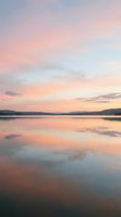 Serene lake at sunset with soft pink and orange sky reflecting on calm water surface, surrounded by distant mountains and hills during golden hour.