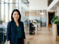 Asian female professional in dark blazer standing in modern office corridor with glass partitions and blurred colleagues, representing corporate business environment and workplace leadership.