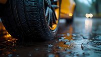 Close-up view of car tire on wet asphalt during rainy evening with bokeh lights in background, reflecting dramatic urban atmosphere and road safety concept.