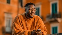 Happy African American man in orange sweater using smartphone outdoors during golden hour sunset with warm lighting and urban architecture background.