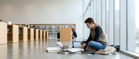 Young man student sitting on library floor with laptop and books studying near large windows in modern academic space for education and research.