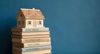 Wooden miniature house model sitting on stack of home design and architecture books against blue background for real estate and interior planning concepts.